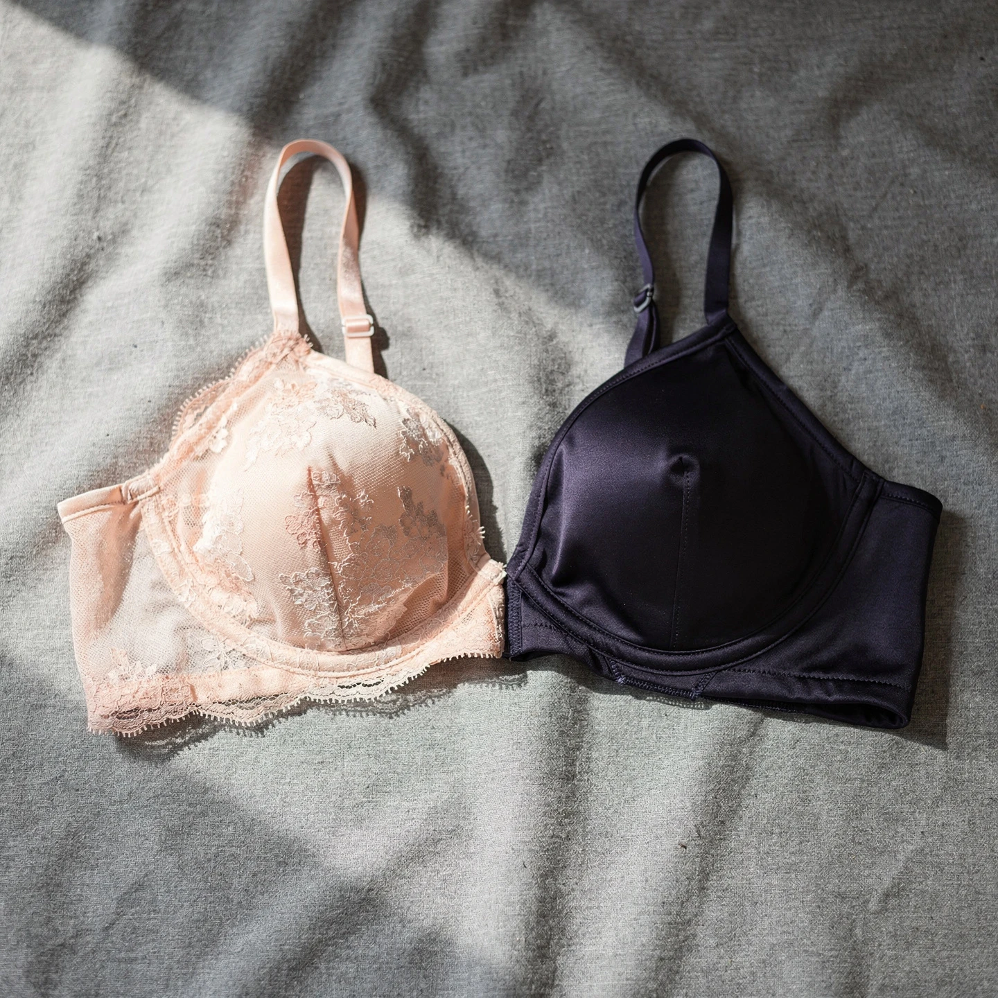 Dwa kontrastujące biustonosze balkonetki: jasny koronkowy i ciemny gładki, ułożone obok siebie na szarej tkaninie, podkreślające różnorodność fasonu, światło dzienne
