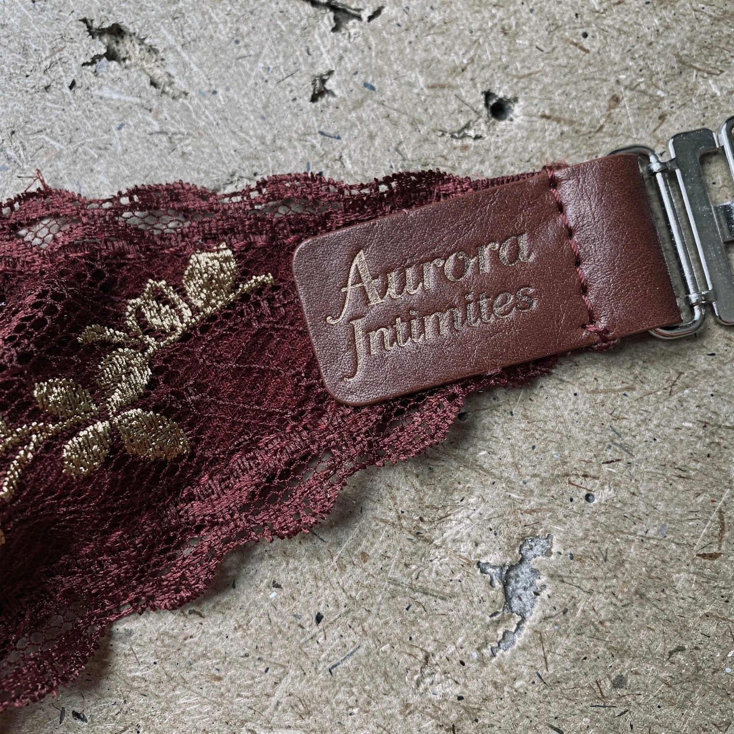 Luksusowy detal zapięcia skrzydełka polskiego biustonosza. Widok zbliżony na zagiętą koronkę w kolorze burgundu z subtelnym złotym brokatem, przyszywaną do skórzanej etykiety z nazwą marki w stonowanym odcieniu. Tło imitujące szorstki karton w odcieniach szarości. Kadr poziomy, ostre skupienie na fakturze koronki i metce.