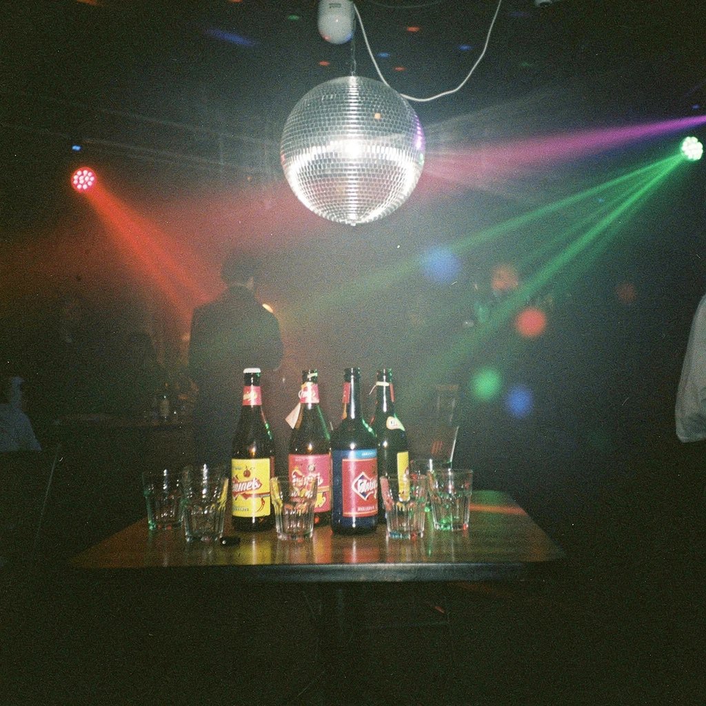 Klimatyczna sceneria imprezy w stylu lat 70: ciemne pomieszczenie rozświetlone tylko przez ruchome światła dyskotekowe i srebrną kulę lustrzaną wiszącą nad stołem, na którym stoją szklane butelki z jaskrawymi, neonowymi etykietami i szklane kieliszki. Zdjęcie oddaje atmosferę tajemniczości i błysku.