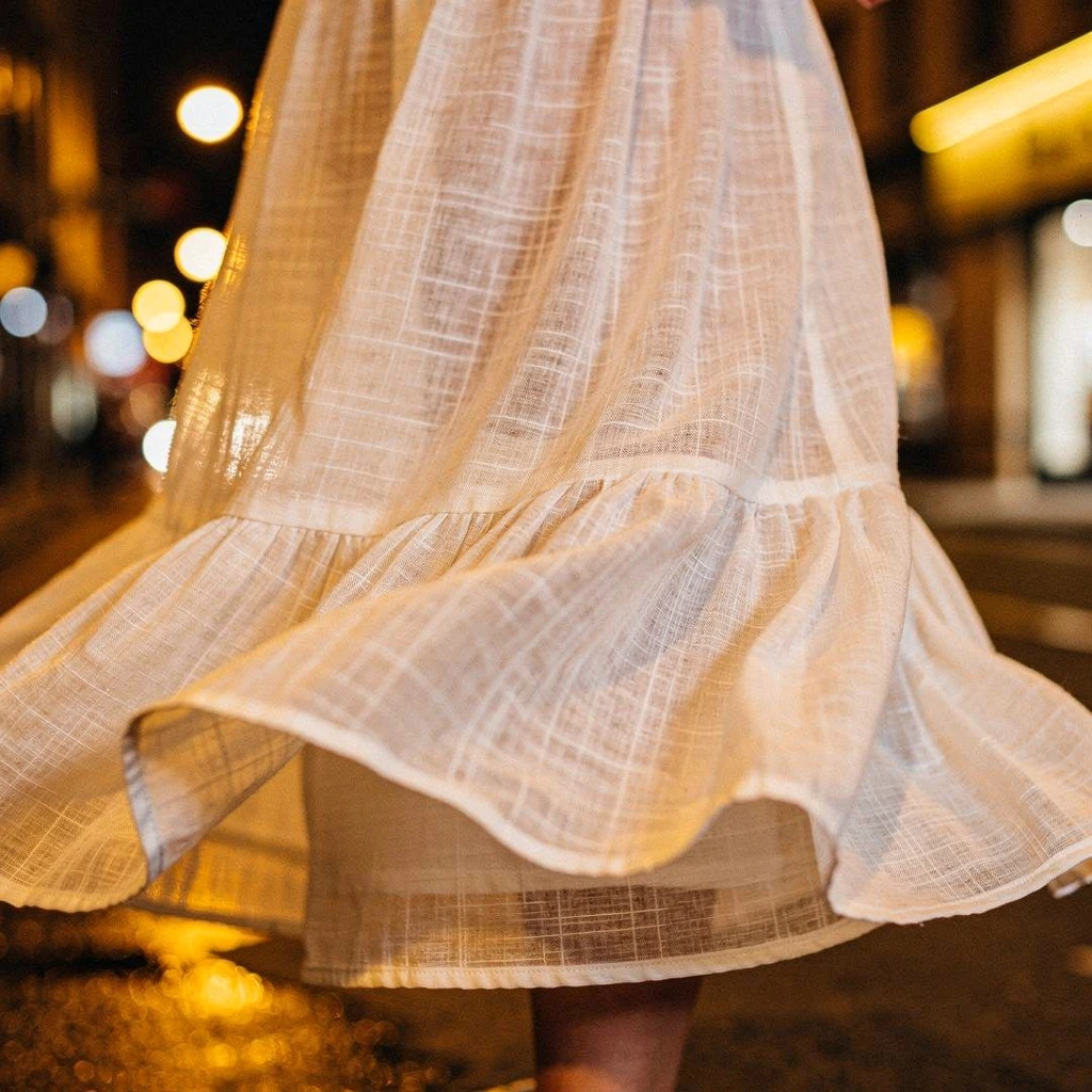 Zwiewna, maxi sukienka w jasnym, naturalnym odcieniu (np. écru), wykonana z lekkiego materiału. Na zdjęciu skupione są na falującym dole sukienki i fakturze tkaniny, w tle rozmyte światła sceny nocą.