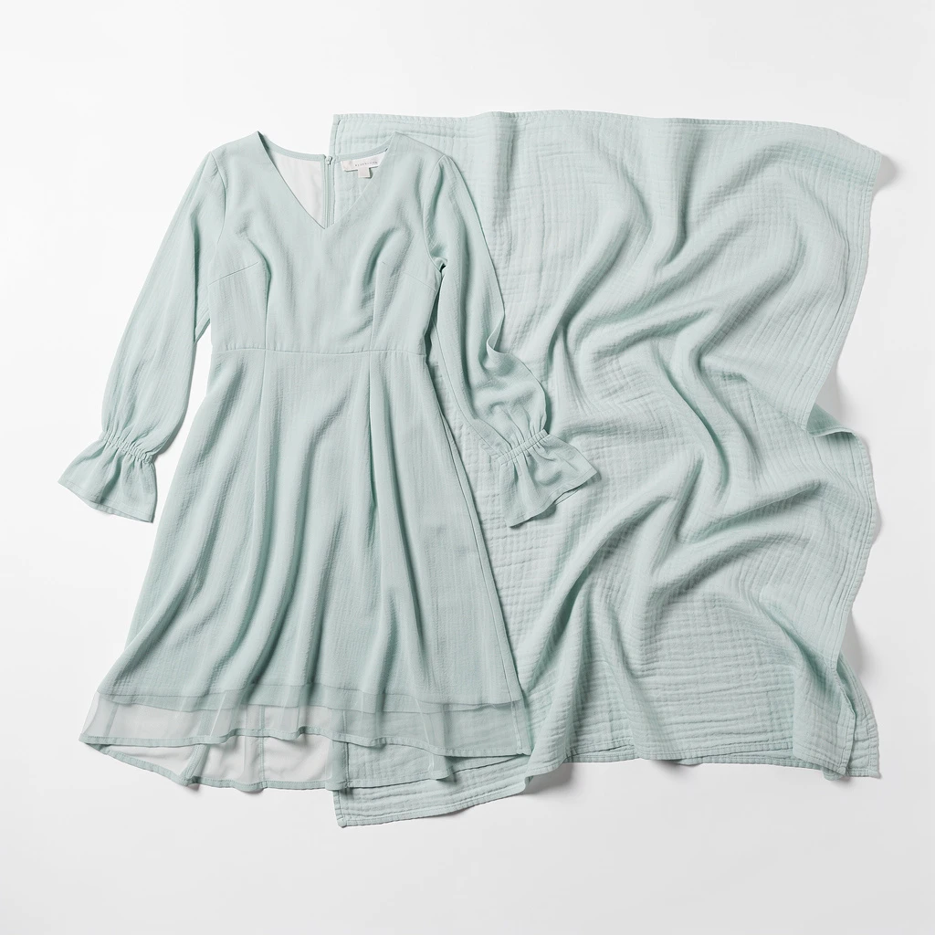 Sukienka w kolorze mięty z delikatną, transparentną narzutką w tym samym odcieniu. Sukienka ma prosty, lekko rozkloszowany krój z długim rękawem i dekoltem w serek. Narzutka wykonana jest z lekkiego, zwiewnego materiału. Zdjęcie pokazuje tylko te dwie części garderoby, ułożone płasko na białym tle, z widoczną fakturą tkanin.