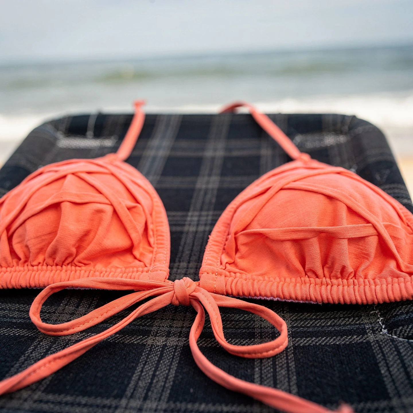 Dwuczęściowy kostium bikini w kolorze koralowym: górą jest trójkątny stanik z drobnymi, poprzecznymi marszczeniami materiału oraz wiązaniem na szyi i plecach, położony na ciemnym plecionym leżaku plażowym na pierwszym planie, w tle delikatnie rozmyte morze.