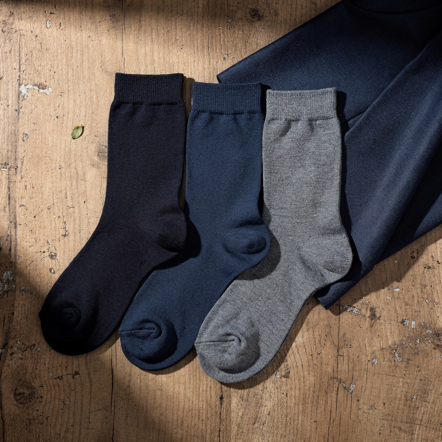 Kompozycja trzech par skarpet w różnych odcieniach granatu ułożonych obok granatowego materiału garniturowego, naturalne drewniane tło, oświetlenie boczne