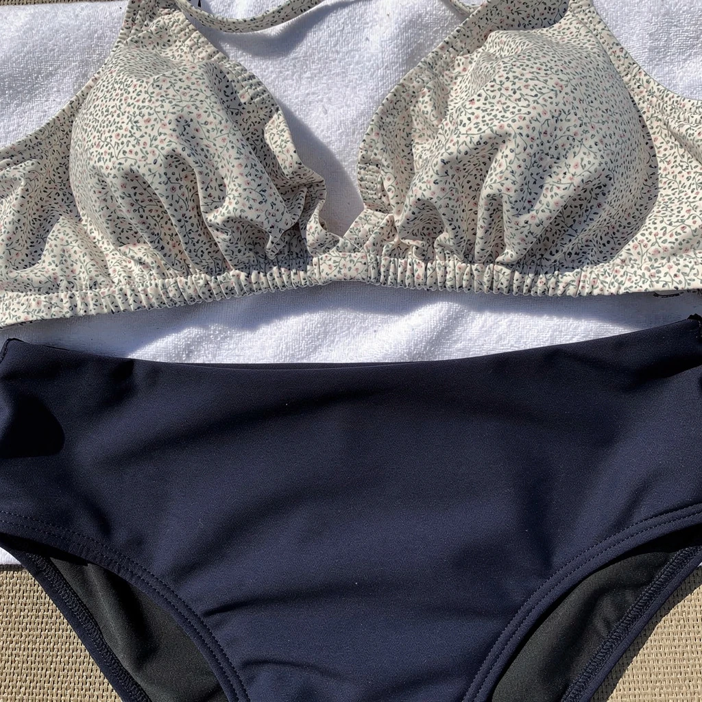 Detal przy makaronie stroju dwuczęściowego: jasny, wzorzysty top bikini (np. w drobne kwiaty) i ciemnogranatowe lub czarne figi z wysokim stanem, widoczne dokładnie w miejscu połączenia. Kadr ukazujący kontrast kolorystyczny między górą a dołem kostiumu. Wybrany kadr plan amerykański, bez osób.